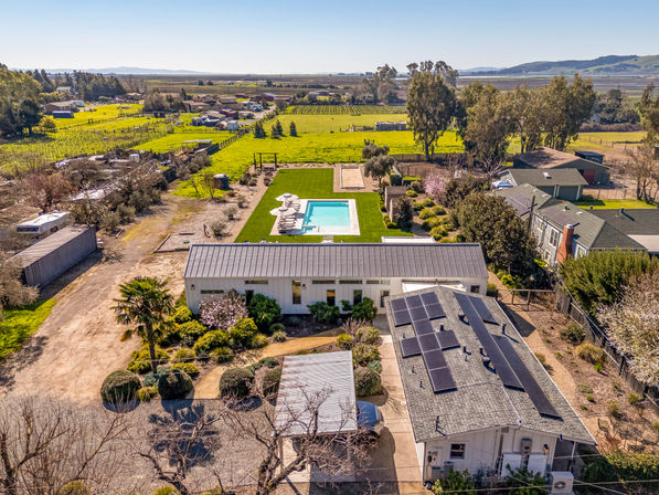 Aerial view of a sunny countryside property with a modern farmhouse, rooftop solar panels, rectangular swimming pool with lounge chairs, manicured lawn and surrounding green farmland and distant hills.