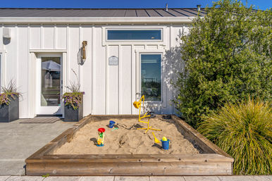 Wooden front-yard sandbox with a playful yellow digger, buckets and shovels in the sand in front of a white modern cottage exterior with planters and green shrubs on a sunny day.