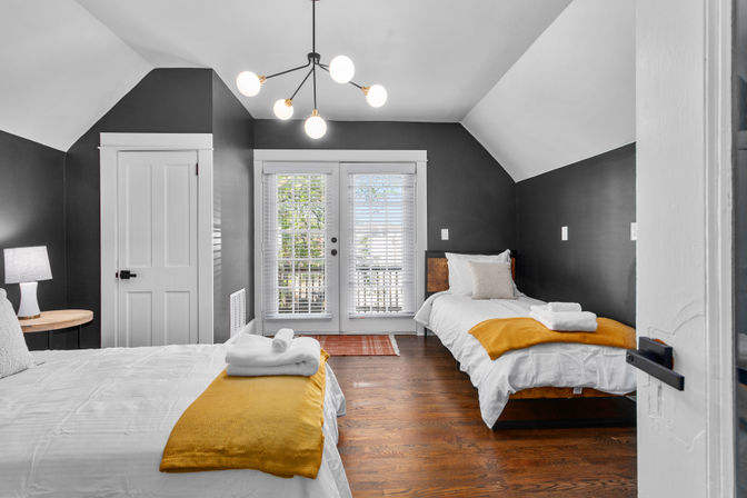 Bright modern attic guest bedroom with two twin beds, dark gray walls, hardwood floors, mustard-yellow throws, white bedding, mid-century chandelier, bedside lamp and French doors with blinds leading to a balcony.