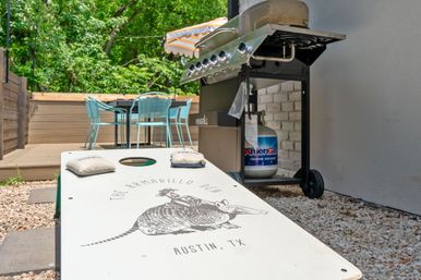 Austin, TX patio with a cornhole board featuring an armadillo graphic, propane grill with tank, and a turquoise outdoor dining set under a striped umbrella.
