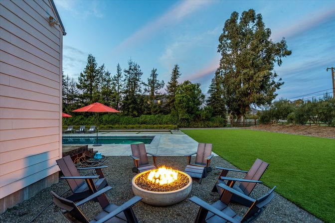 Cozy backyard at dusk with a lit circular fire pit surrounded by Adirondack chairs on a gravel patio, nearby rectangular pool with red umbrellas, manicured lawn and tall trees