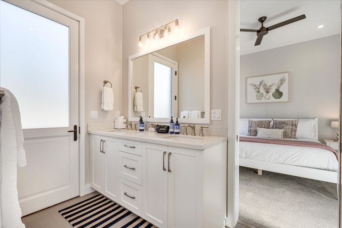 Bright contemporary master bathroom with white double-sink vanity, brass faucets, large mirror, frosted glass door, striped rug, and a view into a cozy bedroom with ceiling fan and botanical wall art.