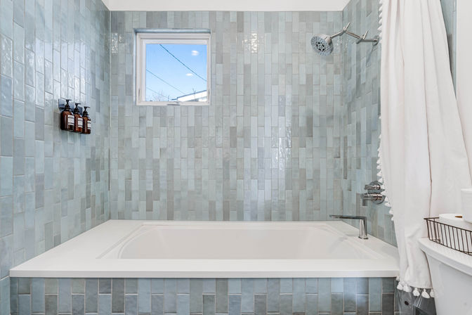 Bright modern bathroom featuring a white soaking tub surrounded by vertical blue-gray subway tiles, chrome showerhead and faucet, three amber wall-mounted soap dispensers, small window with sky view, and a white tassel shower curtain.