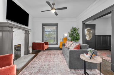 Cozy modern living room interior with a gray sofa, red velvet armchairs, patterned area rug, fireplace with mantel and wall-mounted TV, ceiling fan, large window, indoor plant and side table with books.