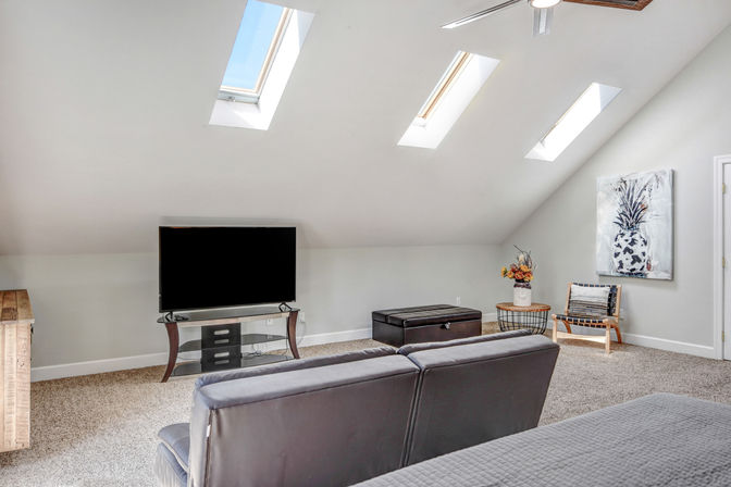 Bright upstairs loft living room with three skylights, flat-screen TV on a glass stand, gray leather sofa, storage ottoman, accent chair, neutral carpet and pineapple wall art