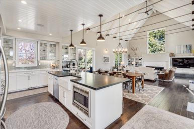 Bright open-concept modern farmhouse kitchen and living room with vaulted white plank ceiling, large island with dark countertop, pendant lights, hardwood floors, dining table, fireplace and tall windows showing trees.