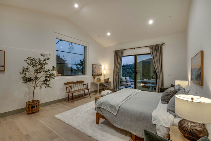 Cozy modern mountain-view bedroom with gray bedding and wooden bed frame, hardwood floors and white rug, sliding glass doors to a patio with outdoor seating, desk by the window, potted tree and warm ambient lighting.