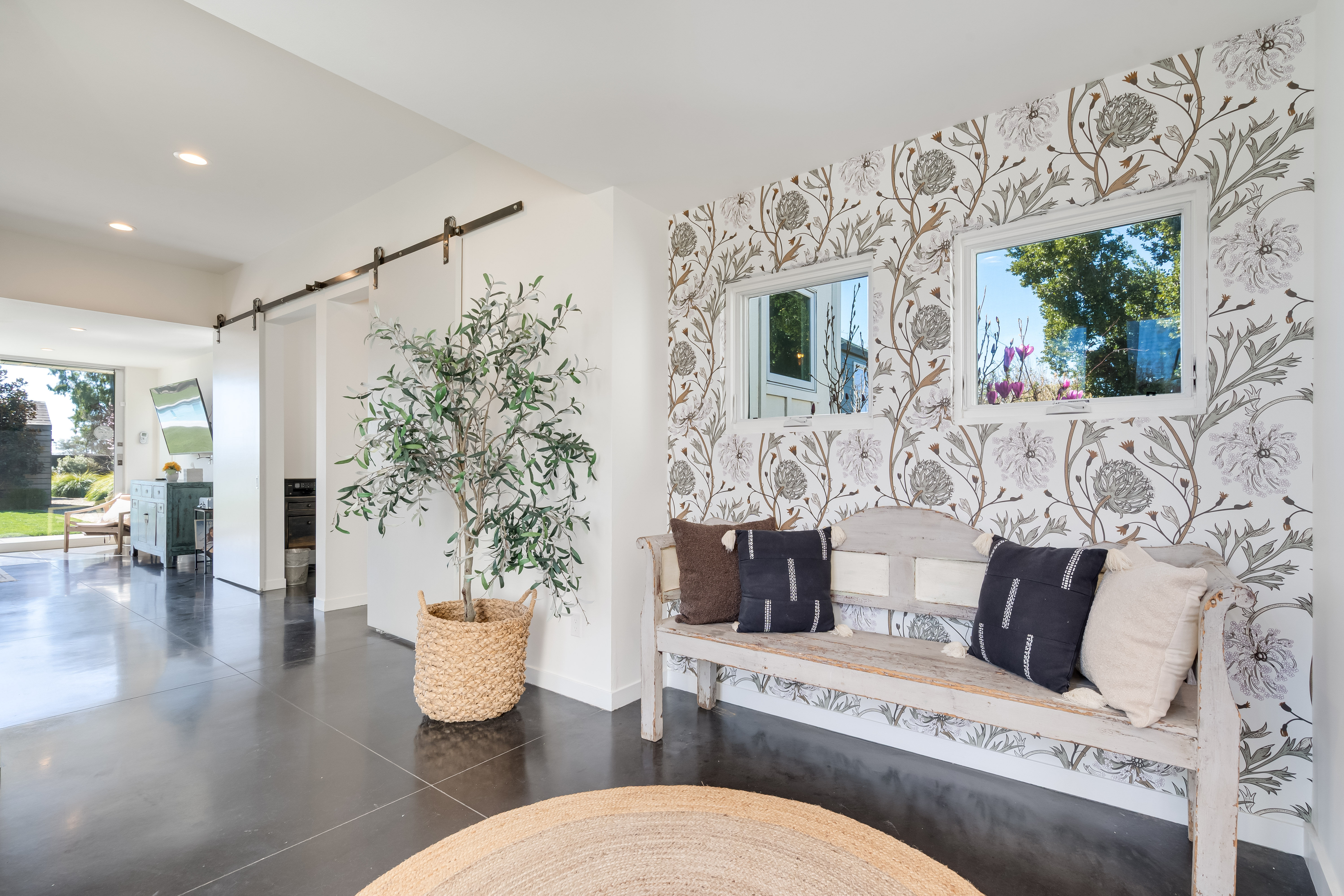Bright modern entryway with floral wallpaper, distressed wooden bench with navy pillows, round woven rug, potted olive tree in a basket, polished concrete floors and barn-style sliding door to an open living space.