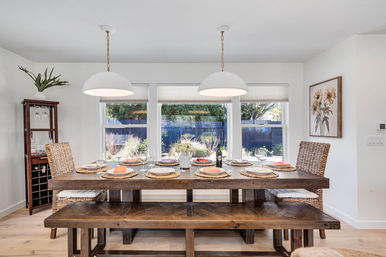 Sunny modern farmhouse dining room with a long rustic wooden table and bench set for six with woven placemats and coral napkins, wicker chairs, white dome pendant lights, floral centerpiece and wine bottle, large windows overlooking a backyard garden.