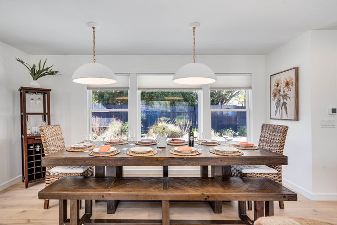 Sunny modern farmhouse dining room with a long rustic wooden table and bench set for six with woven placemats and coral napkins, wicker chairs, white dome pendant lights, floral centerpiece and wine bottle, large windows overlooking a backyard garden.