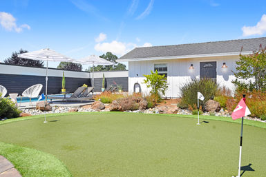 Sunlit suburban backyard with an artificial putting green and numbered flags in the foreground, poolside lounge chairs under striped umbrellas, decorative rock garden and a white pool house beneath a bright blue sky.