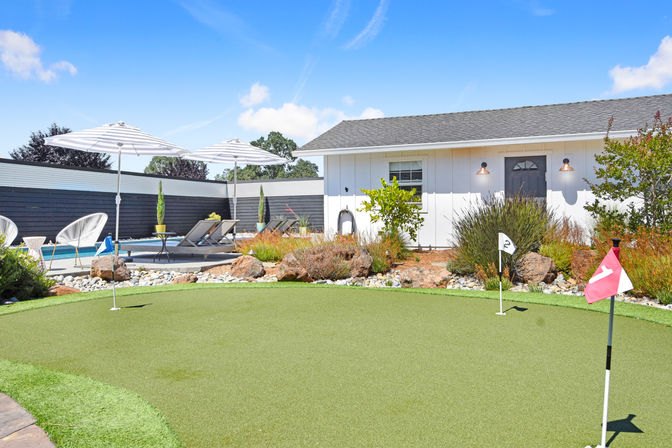 Sunlit suburban backyard with an artificial putting green and numbered flags in the foreground, poolside lounge chairs under striped umbrellas, decorative rock garden and a white pool house beneath a bright blue sky.