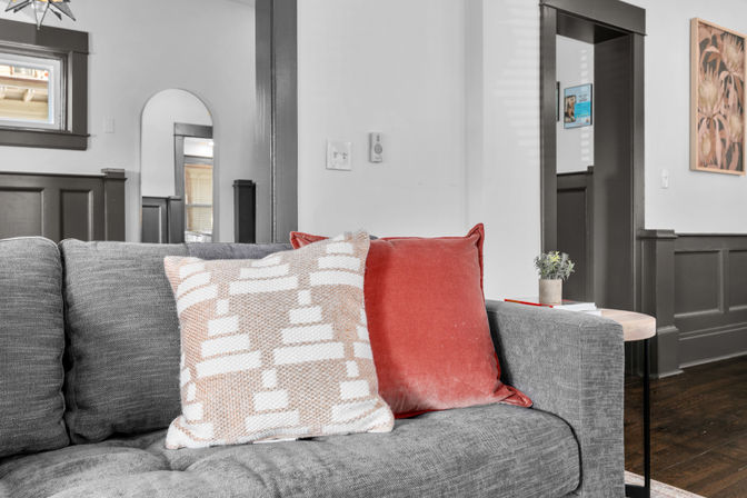 Cozy modern living room interior with a gray fabric sofa, beige-and-white patterned throw pillow and coral velvet cushion, small round side table with a potted plant and books, dark wood trim and hardwood floors.