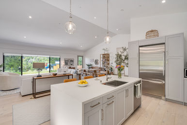 Sunlit open-concept kitchen and living area — white quartz island with undermount sink and bowl of lemons, glass pendant lights, gray shaker cabinets and stainless steel refrigerator, dining and lounge opening to large sliding glass doors with lush backyard view.