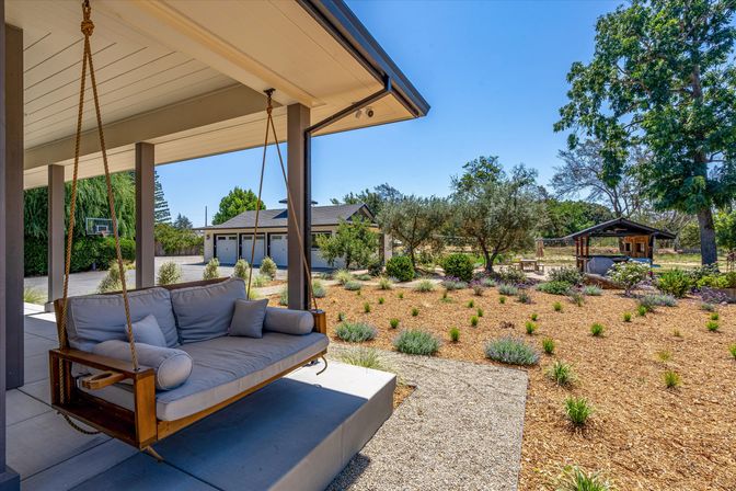 Sunlit covered patio with an inviting cushioned hanging porch swing overlooking a drought-tolerant landscaped backyard with mulch, native shrubs, trees and a detached garage