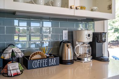 Modern kitchen coffee station on a glossy countertop with a drip coffee maker, single-serve brewer, stainless kettle, tea packet rack, paper filters and a 'coffee bar' organizer against a blue subway tile backsplash and white cabinets.