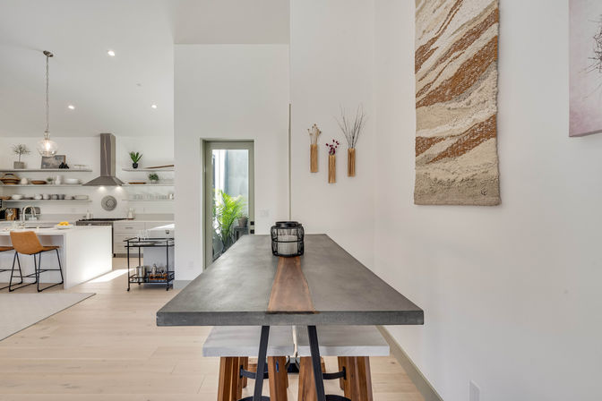 Bright, airy open-concept modern kitchen and dining area with a concrete-and-wood counter table, wooden bar stools, white cabinetry, stainless range hood, pendant light, woven wall tapestry and potted plants.