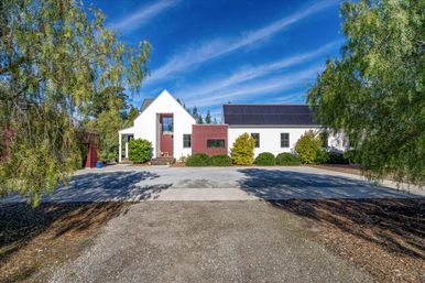 Modern white farmhouse with red accent panels and rooftop solar panels, landscaped shrubs and gravel driveway under a bright blue sky with wispy clouds, framed by leafy trees.