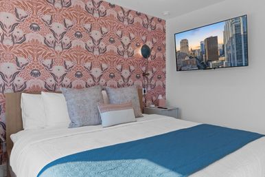 Boutique-style bedroom with playful pink floral wallpaper, king bed dressed in white linens and a blue quilted throw, patterned pillows, wall-mounted TV showing a downtown skyline, and a bedside lamp and nightstand.