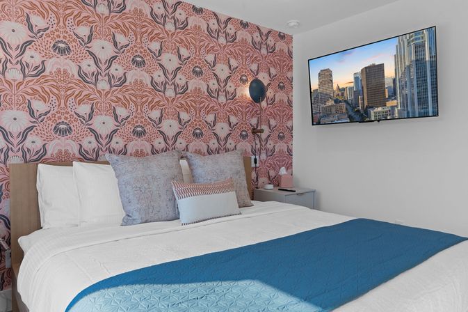 Boutique-style bedroom with playful pink floral wallpaper, king bed dressed in white linens and a blue quilted throw, patterned pillows, wall-mounted TV showing a downtown skyline, and a bedside lamp and nightstand.