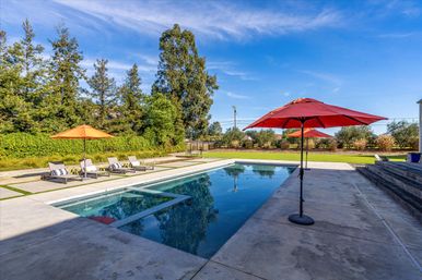 Sunny backyard oasis with a rectangular in-ground pool, concrete patio, red umbrellas and lounge chairs beside a manicured lawn and tall trees.