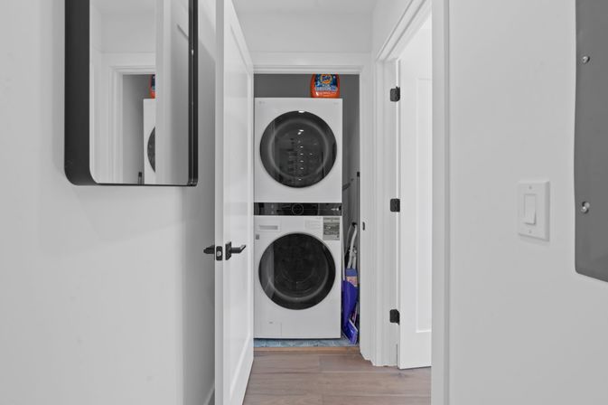 Peek into a compact hallway laundry closet with stacked front-loading washer and dryer, a laundry detergent jug on top and cleaning tools tucked beside the units.
