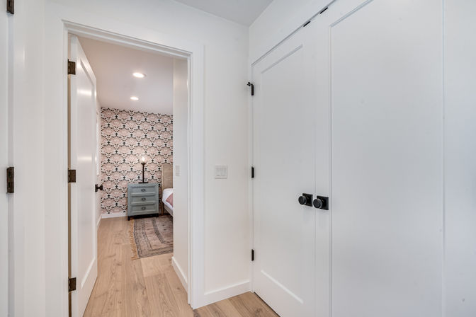 Bright white hallway with wood floors and black-doorknob double closet doors opening to a bedroom featuring scallop-patterned wallpaper, a teal nightstand, floor lamp and area rug.