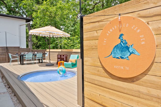 Sunny Austin backyard deck with a small circular plunge pool, striped patio umbrella, dining table and colorful lounge chair, plus a round orange sign showing a whimsical armadillo illustration.