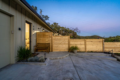 Twilight view of a modern home entry and concrete driveway featuring a wooden bench, horizontal cedar privacy fence, potted plants, and rolling hillside backdrop.