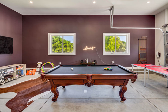 Home game room in a converted garage featuring an ornate wooden pool table with black felt, ping-pong table, neon “cheers” sign, windows and concrete floor.
