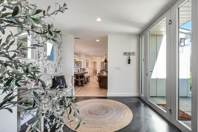 Bright modern foyer with floor-to-ceiling glass doors, round braided jute rug, rustic white bench with black throw pillows, botanical wallpaper and olive-branch plant, opening into a dining and living area