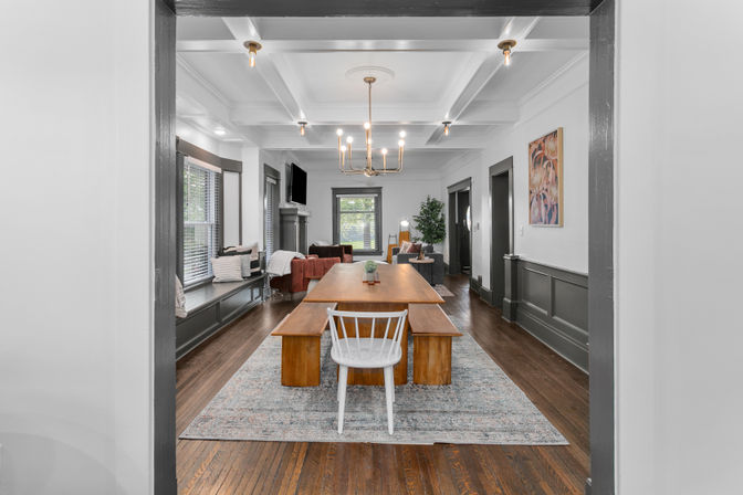 Open-plan dining and living room with a long wooden table, bench and white chair on a patterned rug, coffered ceiling with brass chandelier, hardwood floors, built-in window seat and seating area with fireplace in the background.