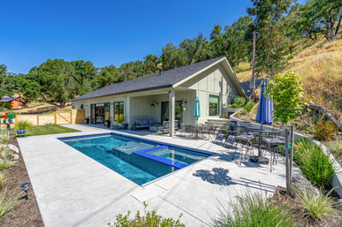 Sunny modern hillside backyard with rectangular in-ground pool and attached spa, concrete patio with lounge chairs, dining table and umbrellas, seating area by a contemporary single-story home, oak trees and grassy slope in the background