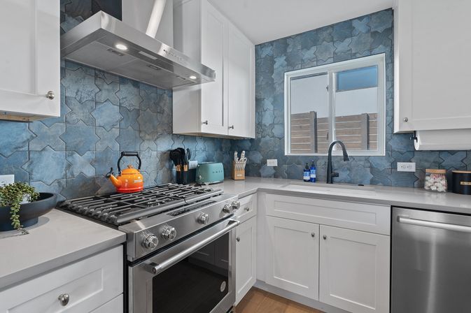 Bright modern residential kitchen with blue star-pattern tile backsplash, stainless-steel gas range and hood, white shaker cabinets, black faucet, teal toaster and orange kettle.