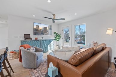 Bright, cozy modern living room in a suburban home with a brown leather sofa, gray swivel chair, patterned rug, teal geometric console under a wall TV, ceiling fan, potted plant, and large windows overlooking a tree-lined street.