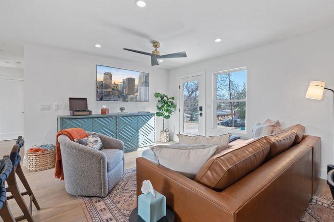 Bright, cozy modern living room in a suburban home with a brown leather sofa, gray swivel chair, patterned rug, teal geometric console under a wall TV, ceiling fan, potted plant, and large windows overlooking a tree-lined street.