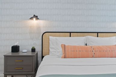 Minimal modern bedroom with rattan headboard, white bedding and coral patterned lumbar pillow, gray nightstand holding a tissue box, small succulent and compact smart device, and a lit black wall sconce against textured gray wallpaper.