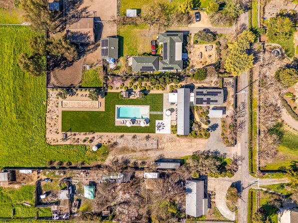 Aerial drone view of a sunlit rural property: rectangular swimming pool with umbrellas on a manicured lawn, patio dining area, sand court, solar panels on multiple roofs, outbuildings, garden plots and a tree-lined access road.