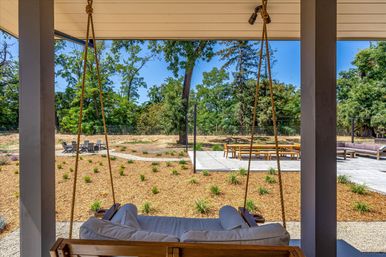 Covered porch with a rope-hung wooden swing facing a landscaped backyard with a concrete patio, long outdoor dining table, lounge seating and a fire-pit seating area among mature trees under a clear blue sky.