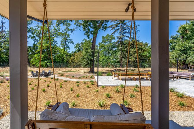 Covered porch with a rope-hung wooden swing facing a landscaped backyard with a concrete patio, long outdoor dining table, lounge seating and a fire-pit seating area among mature trees under a clear blue sky.