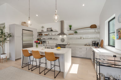 Sunlit modern kitchen with white waterfall island, three tan leather bar stools, glass pendant lights, stainless steel refrigerator and range hood, open white shelves with dishes, hardwood floors and leafy plant.