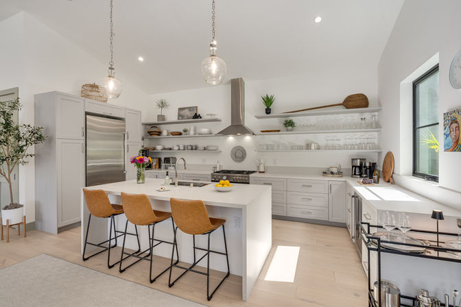 Sunlit modern kitchen with white waterfall island, three tan leather bar stools, glass pendant lights, stainless steel refrigerator and range hood, open white shelves with dishes, hardwood floors and leafy plant.