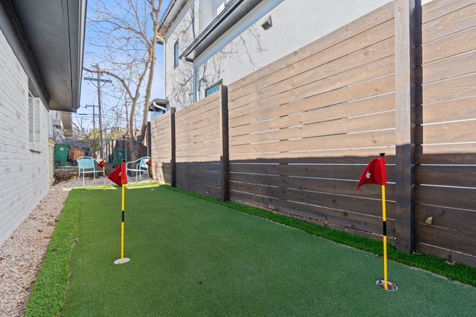 Narrow urban townhouse backyard with a mini artificial putting green, three red-flag holes, wooden privacy fence and two patio chairs on a sunny day.