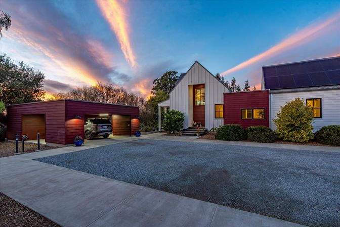 Modern farmhouse-style home with steep white gable and red detached garage revealing an SUV, solar panels on the roof, gravel driveway and dramatic streaked clouds bathed in sunset glow.