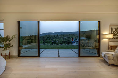 Modern living room with wide sliding glass doors opening to a grassy patio and tree-covered hills at dusk, light wood floors, a potted plant on the left and a cozy armchair with lamp on the right.
