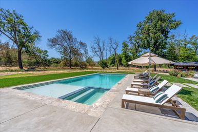 Sunny countryside backyard outdoor swimming pool with built-in spa, stone deck and row of wooden lounge chairs with striped pillows and umbrella, grassy lawn and mature trees under a clear blue sky.