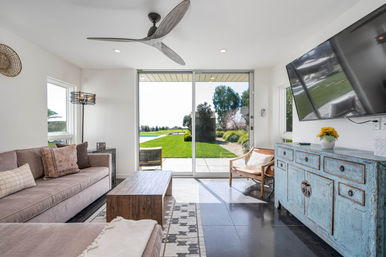 Bright modern living room with large sliding glass doors opening to a grassy backyard and pool, neutral sofa, wooden coffee table, distressed blue sideboard and wall-mounted TV.