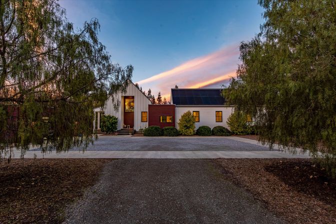 Modern farmhouse with white and wood siding, solar panels on the roof, warm interior lights and a gravel driveway framed by leafy trees under a pink sunset sky.
