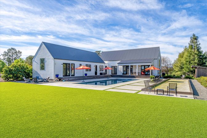 Modern white farmhouse with gabled metal roofs, backyard swimming pool and patio with red umbrellas, lounge chairs and a manicured green lawn under a blue sky.