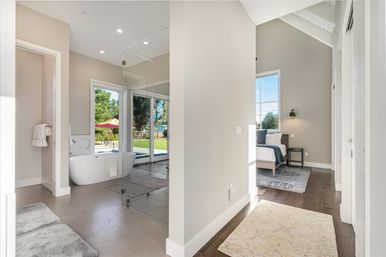 Sunlit modern master suite with open-concept bathroom—freestanding tub and frameless glass shower—sliding glass doors to a green backyard with pool and red umbrella, adjacent vaulted bedroom with hardwood floors and neutral decor.